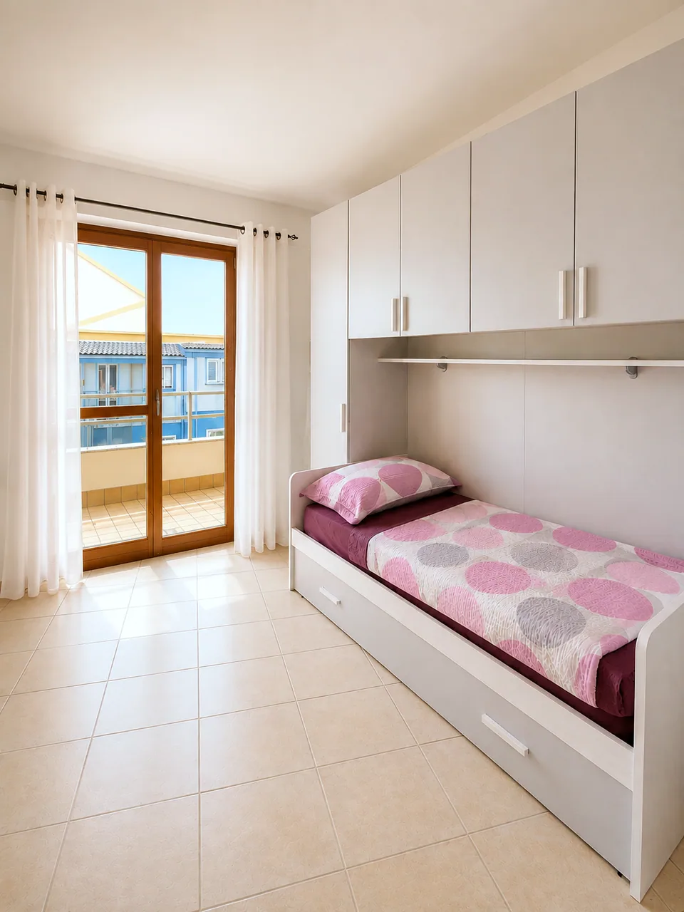 Second bedroom single bed beside sheer-curtained balcony doors in full sunlight