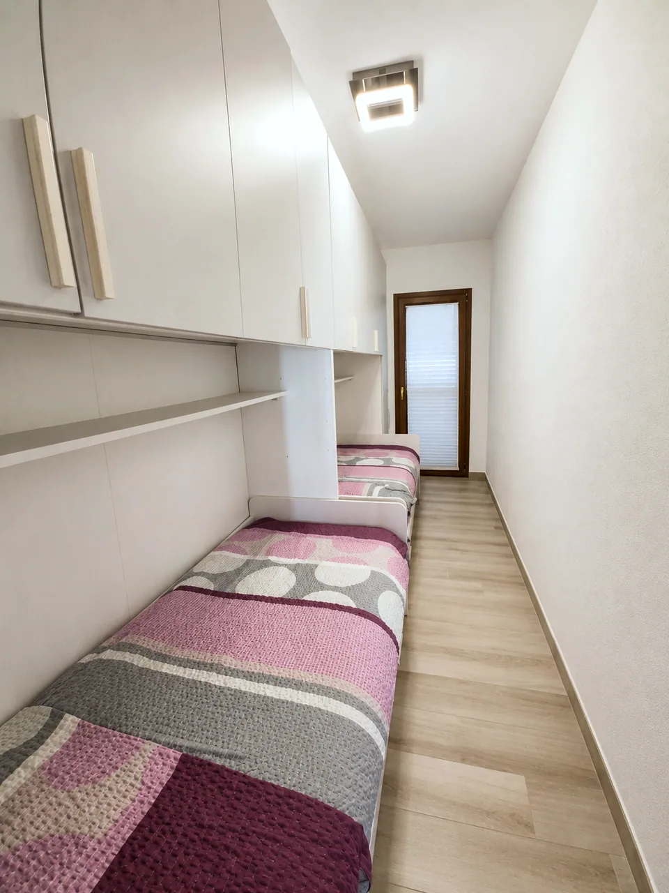 View along the second bedroom at Il Paradiso past the twin beds and light oak flooring towards the shuttered balcony door