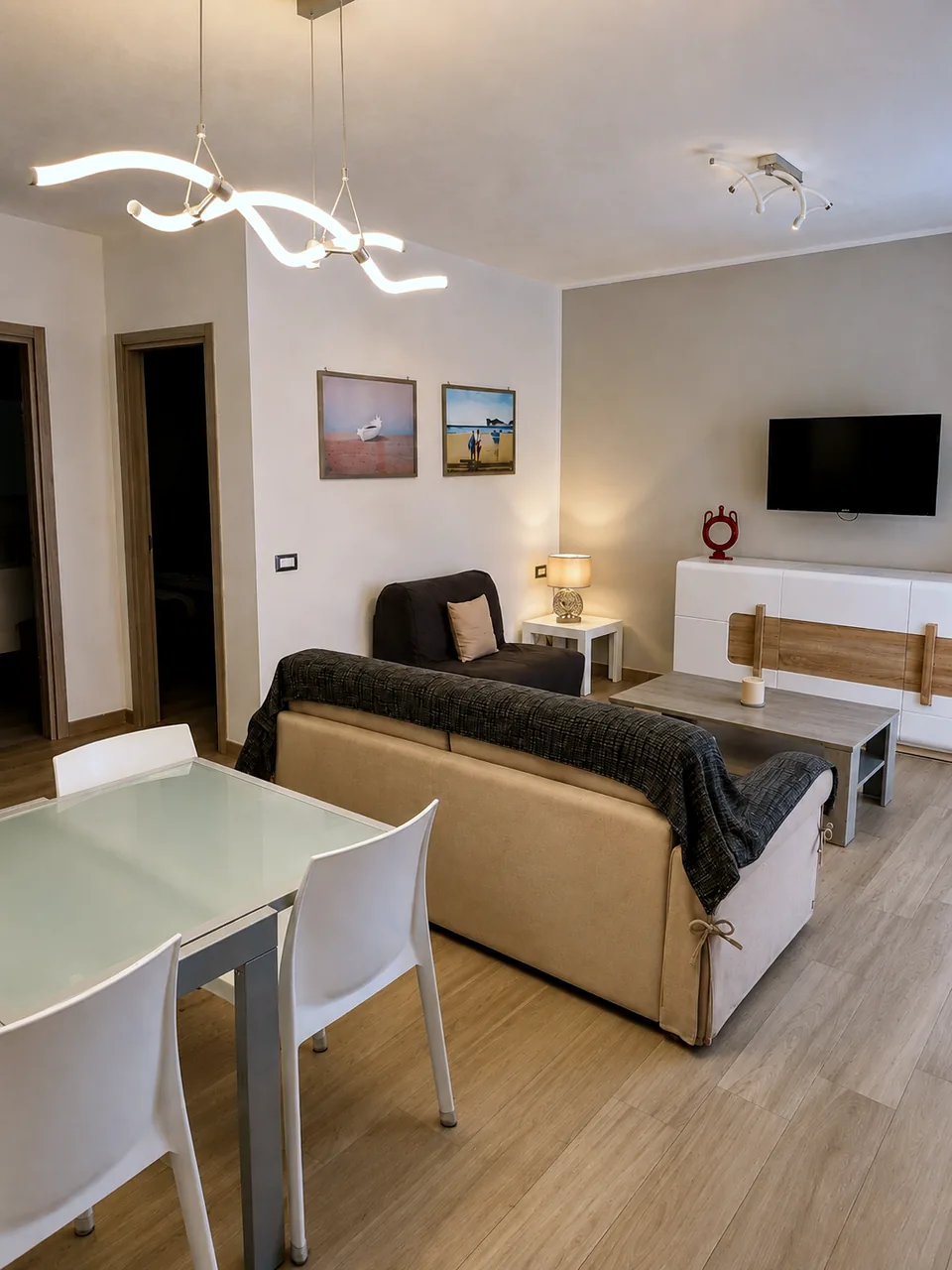 Evening view of the Il Paradiso lounge with beige sofa bed, armchair, table lamp, coastal artwork and a wall-mounted flat-screen TV