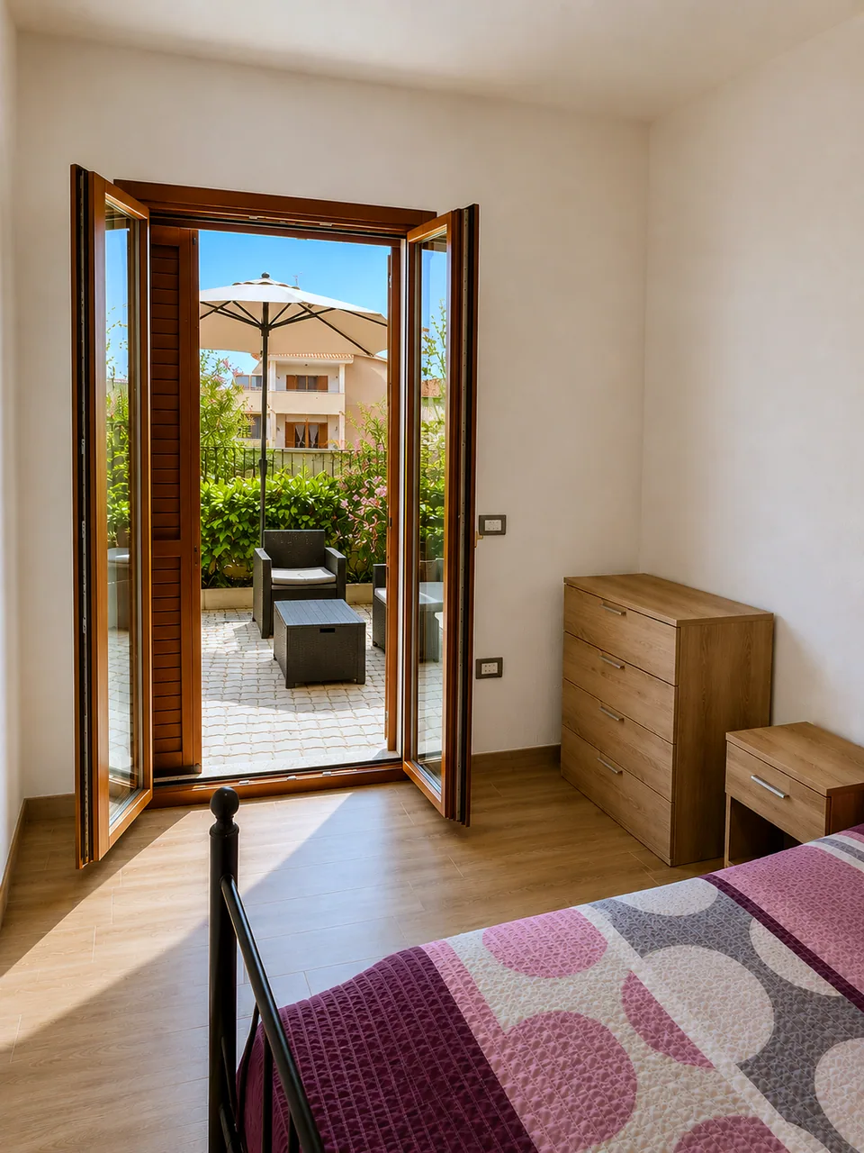 Main bedroom at Il Paradiso with wooden French doors open onto the sunlit private terrace with parasol, rattan seating and flowering shrubs