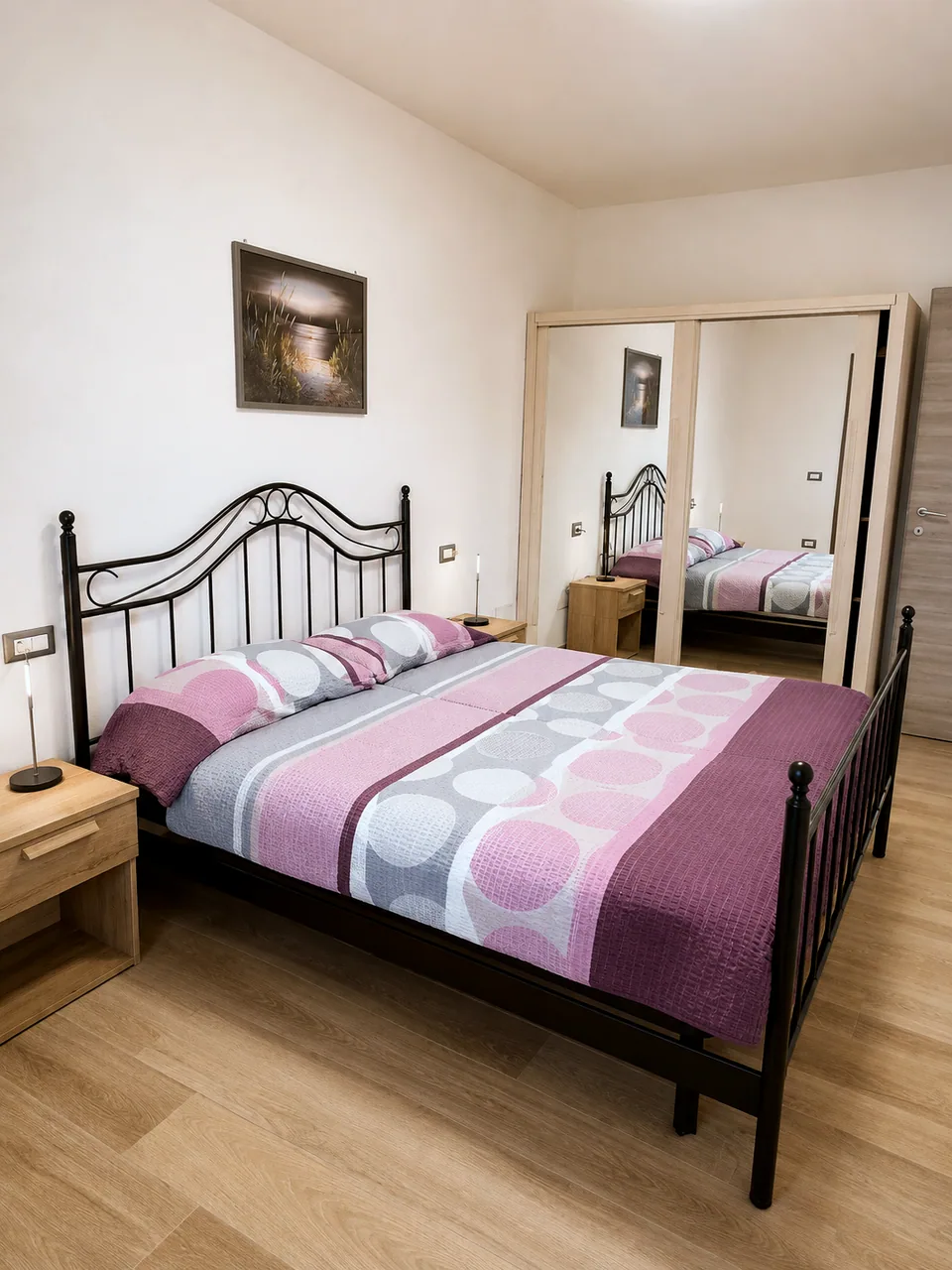 Main bedroom at Il Paradiso in warm evening lamplight showing the large mirrored oak wardrobe, iron bed frame and pink and grey bedspread