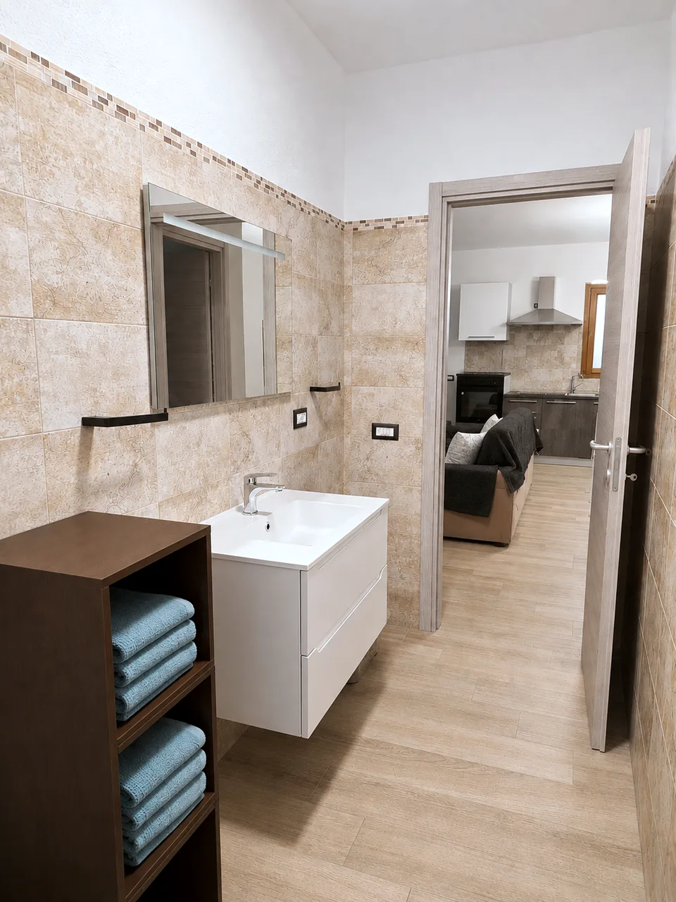 Il Paradiso bathroom vanity with illuminated mirror, folded turquoise towels on a dark wood shelf unit and the open door looking through to the lounge and kitchen