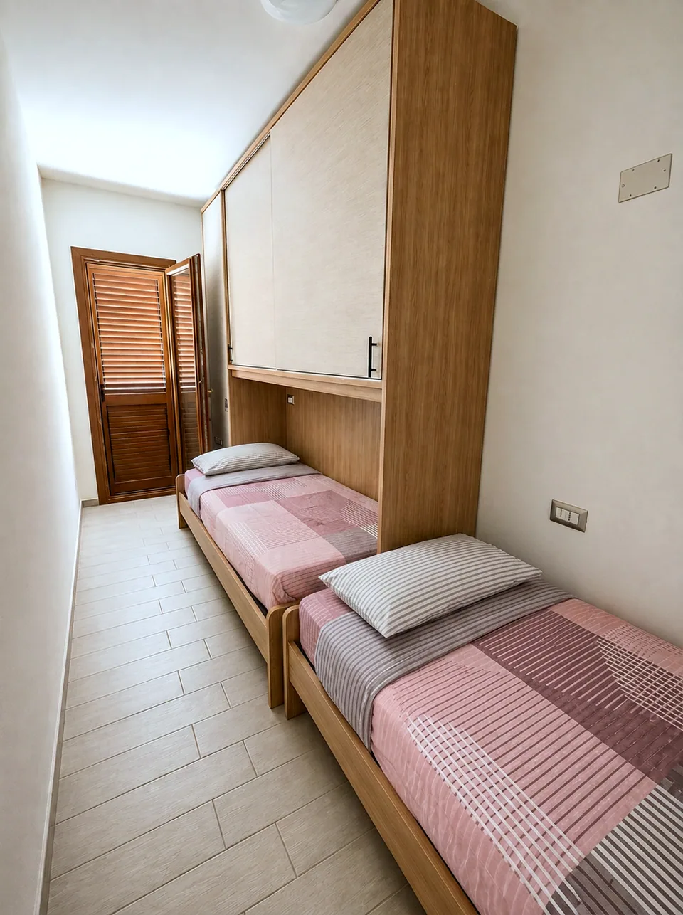 Twin bedroom at La Dolce Vita looking towards the louvred wooden balcony door, with both single beds made up in pink and grey linen