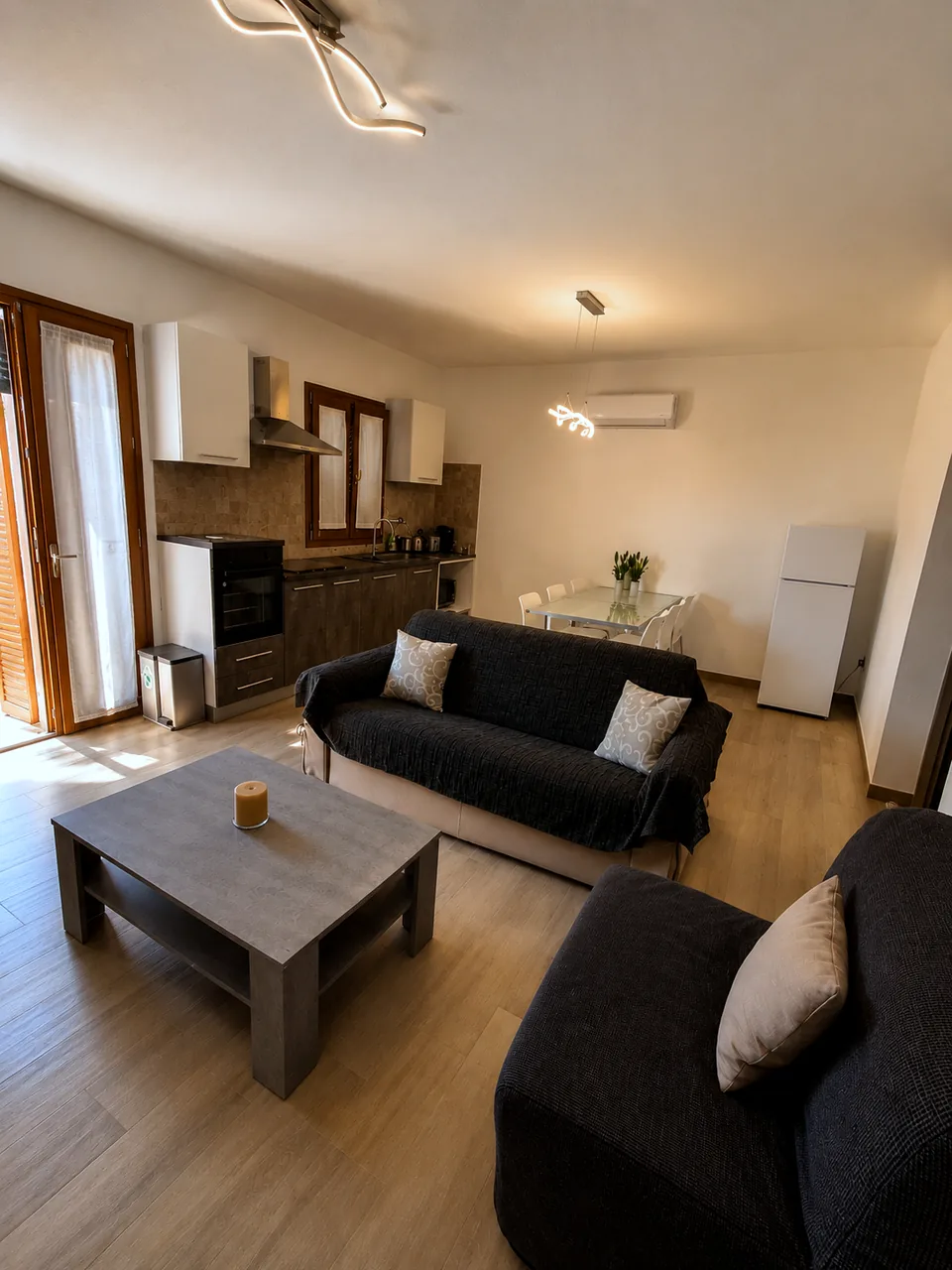 Open-plan lounge and kitchen at Il Paradiso in warm evening light with charcoal sofas, grey coffee table and the balcony door open