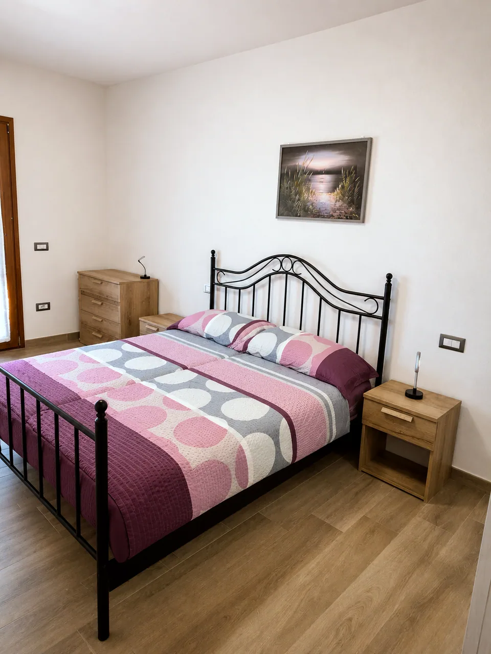 Main bedroom at Il Paradiso in daylight with a black wrought-iron double bed, pink and grey bedding, oak chest of drawers and bedside table and a coastal sunset print