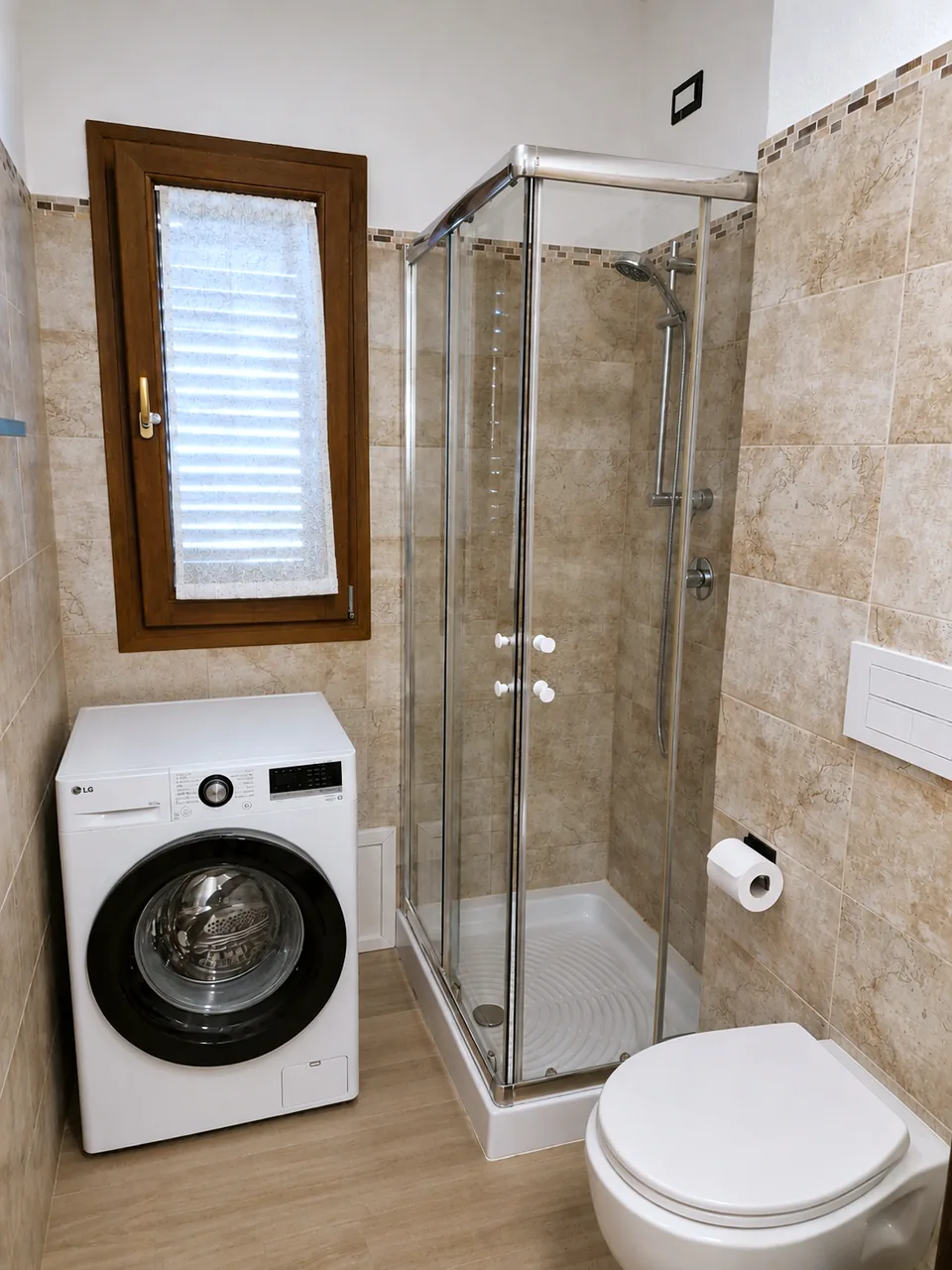 Corner shower with glass sliding doors, handheld shower, wall-hung toilet and washing machine beneath the wooden-framed bathroom window at Il Paradiso