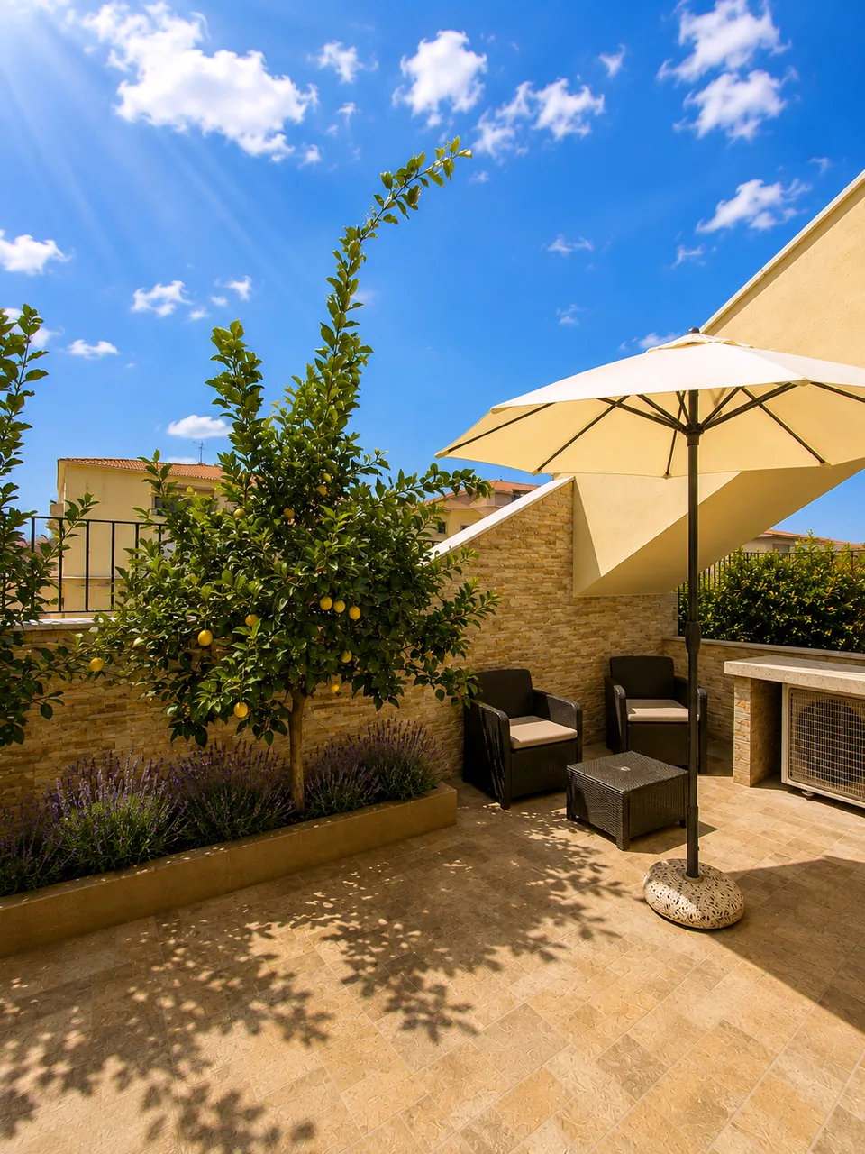 Sunny bedroom-side terrace at La Dolce Vita with a lemon tree in fruit, lavender bed, cream parasol and dark rattan armchairs on travertine paving