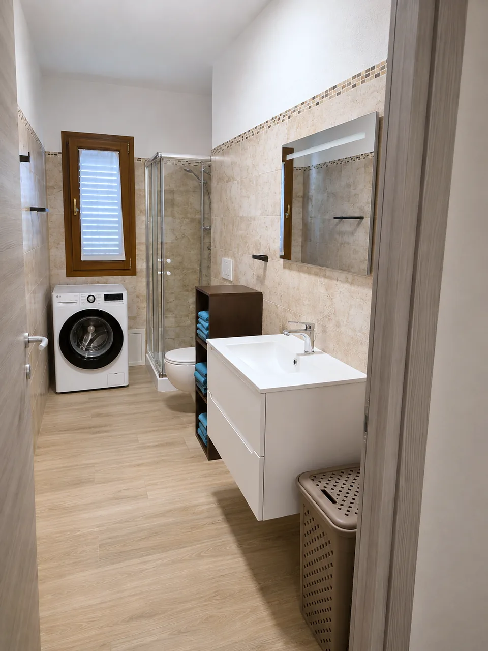 Bathroom at Il Paradiso with beige marble-effect tiling, white wall-hung vanity, illuminated mirror, corner shower, toilet, LG washing machine and light oak-look floor
