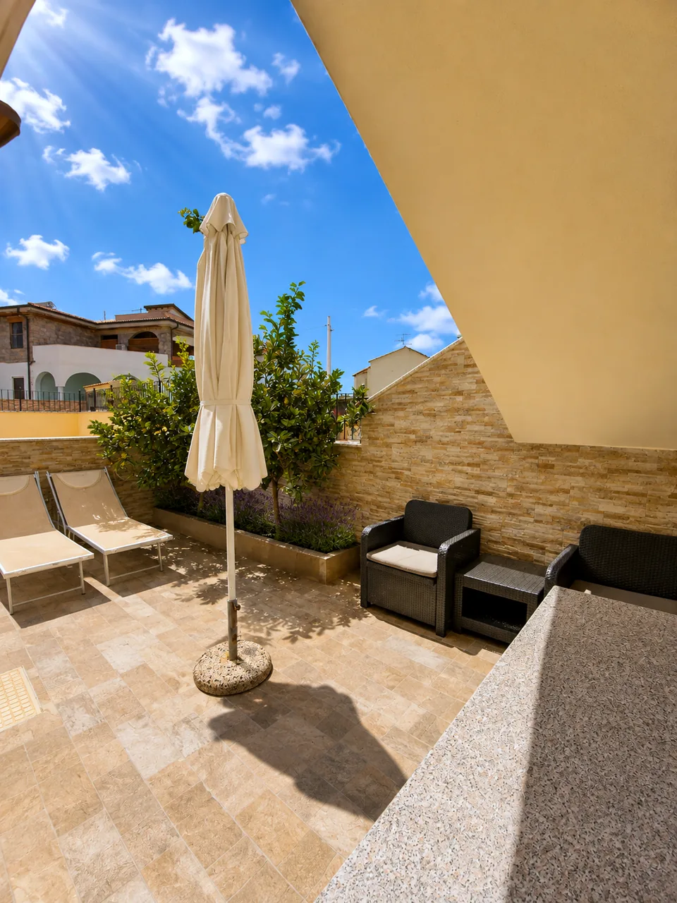 Bedroom-side terrace at La Dolce Vita with two cream sun loungers, rattan lounge seating and a stone-clad wall shaded by citrus trees