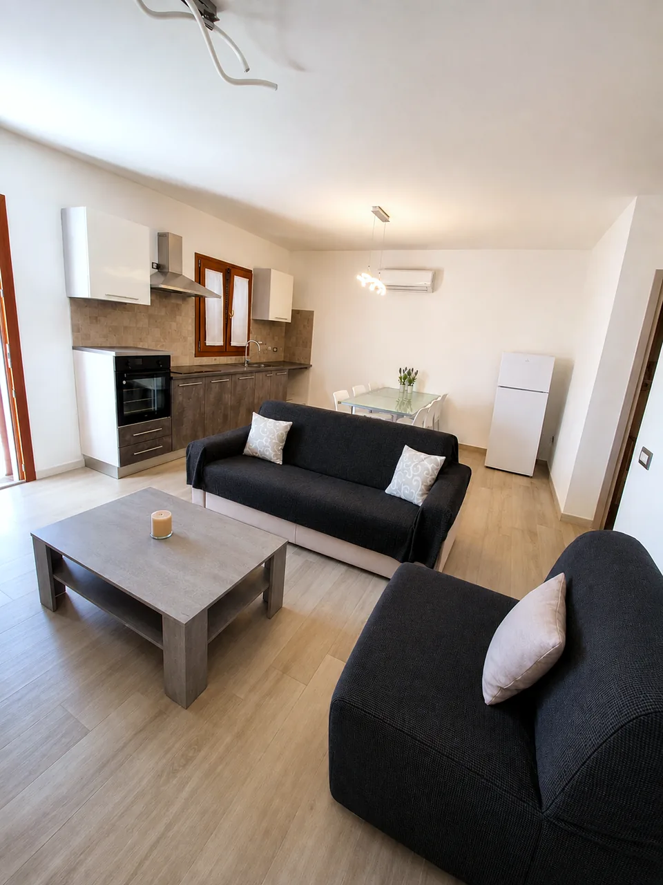 Daylight view across the open-plan living area at Il Paradiso showing two charcoal sofas, a grey concrete coffee table and the kitchen with oven and fridge