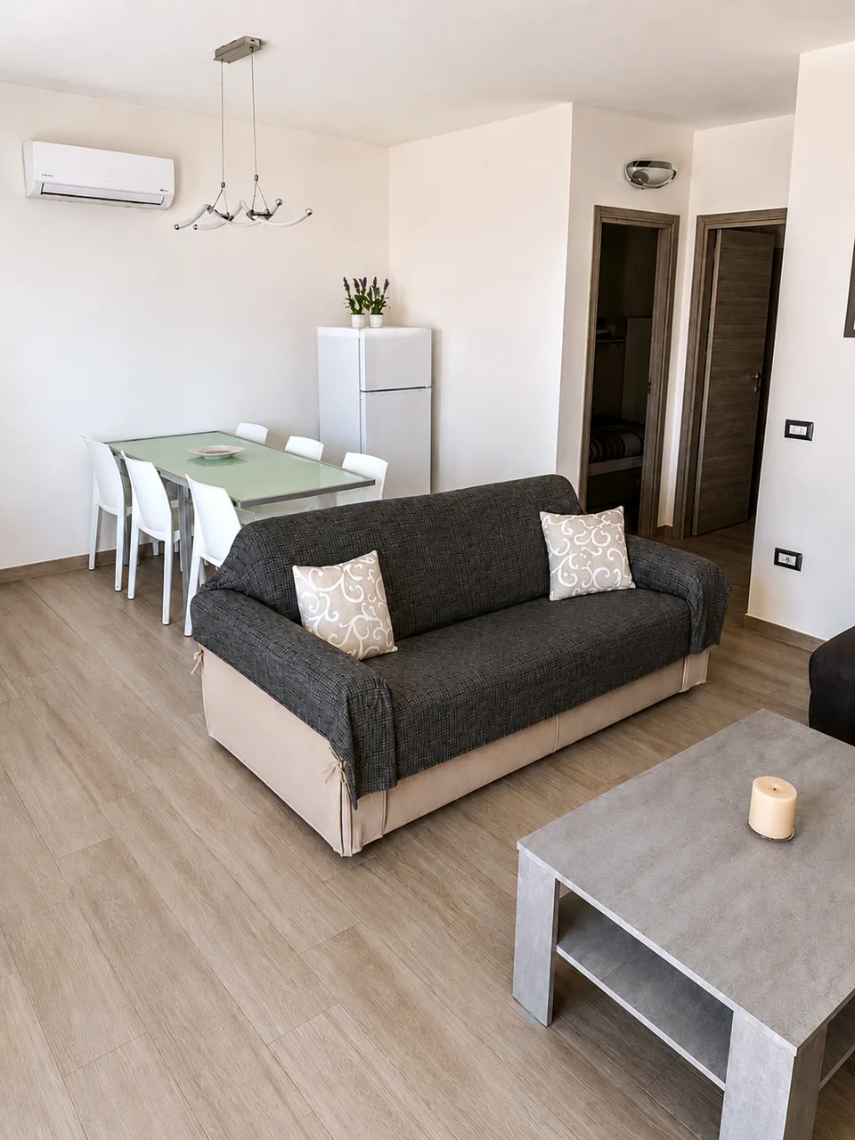 Open-plan lounge and dining room at Il Paradiso in daylight with a charcoal-covered sofa bed on a beige base, glass dining table with white chairs and air conditioning