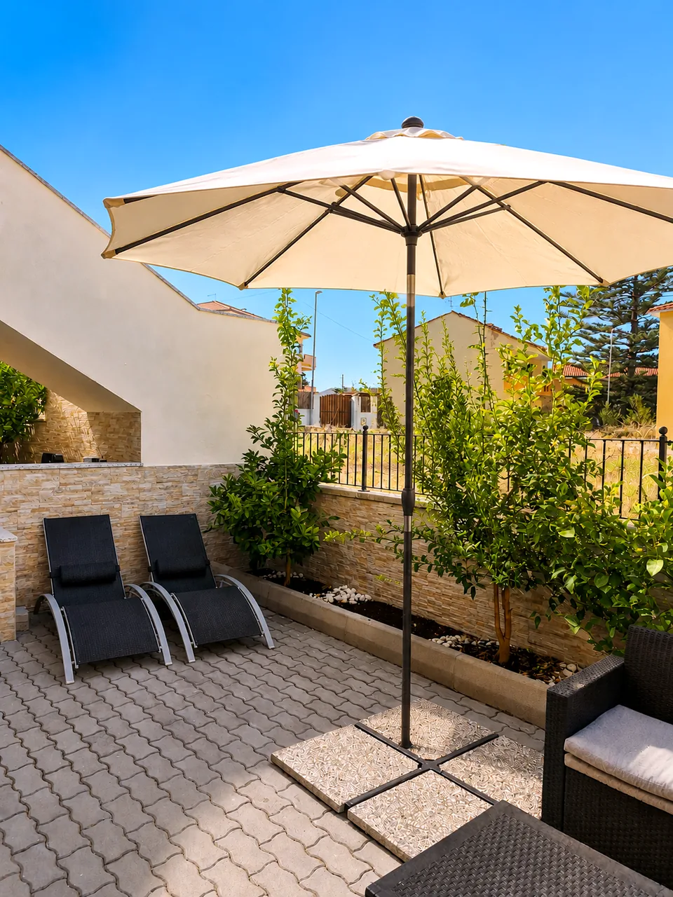 Two black sun loungers on the rear terrace at Il Paradiso beside citrus trees, stone-clad wall and a large cream parasol under a clear blue sky